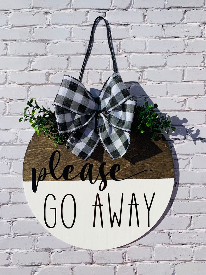 May include: A round wooden door hanger with a black and white checkered bow, green foliage, and the text "Please Go Away" in black lettering.
