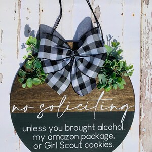 Puede incluir: Un colgador de puerta redondo de madera con un lazo a cuadros blanco y negro y follaje verde. El letrero dice "no soliciting unless you brought alcohol, my amazon package, or Girl Scout cookies."