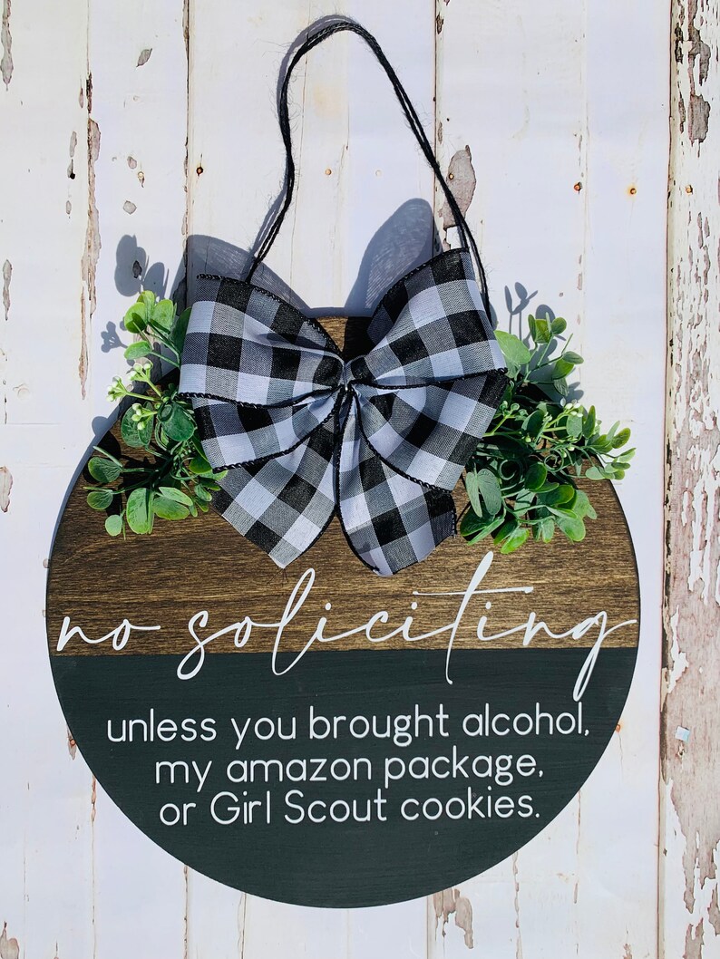 Puede incluir: Un colgador de puerta redondo de madera con un lazo a cuadros blanco y negro y follaje verde. El letrero dice "no soliciting unless you brought alcohol, my amazon package, or Girl Scout cookies."