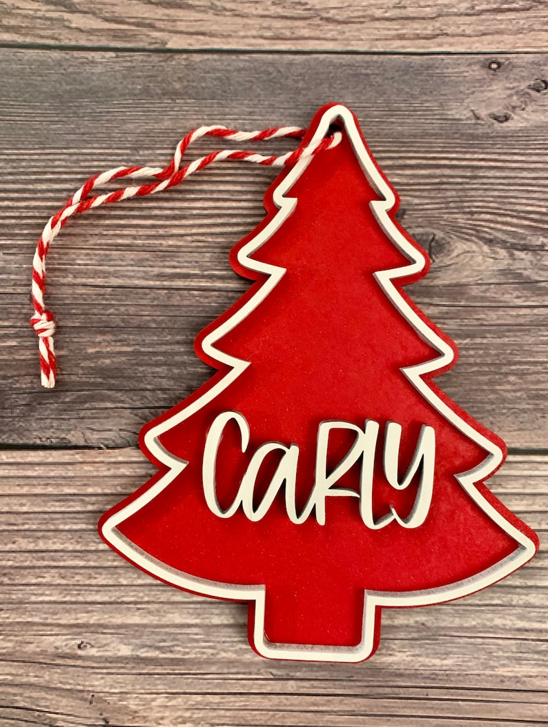 K&ouml;nnte beinhalten: Roter Weihnachtsbaum-Anh&auml;nger mit wei&szlig;em Rand und dem Namen "Carly" in wei&szlig; auf der Vorderseite.