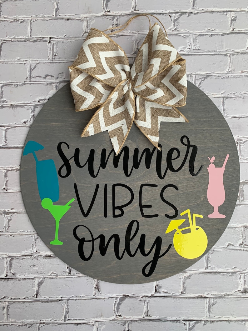 K&ouml;nnte beinhalten: Runder, grauer Holzschild mit dem Schriftzug "Summer Vibes Only" in schwarzer Schrift. Es zeigt farbenfrohe Cocktail-Illustrationen und eine dekorative Schleife mit Zickzackmuster. Ein Juteseil ist zum Aufh&auml;ngen befestigt.