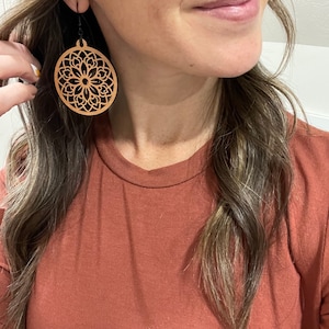 May include: A pair of large, round wooden earrings with a delicate, intricate laser-cut design. The earrings are a light brown color and have a black hook.