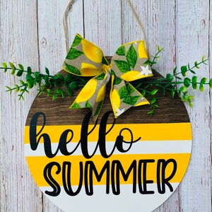 May include: A round wooden sign with a yellow and white striped background and the text "hello summer" in black. A yellow and green lemon print bow is attached to the top of the sign.