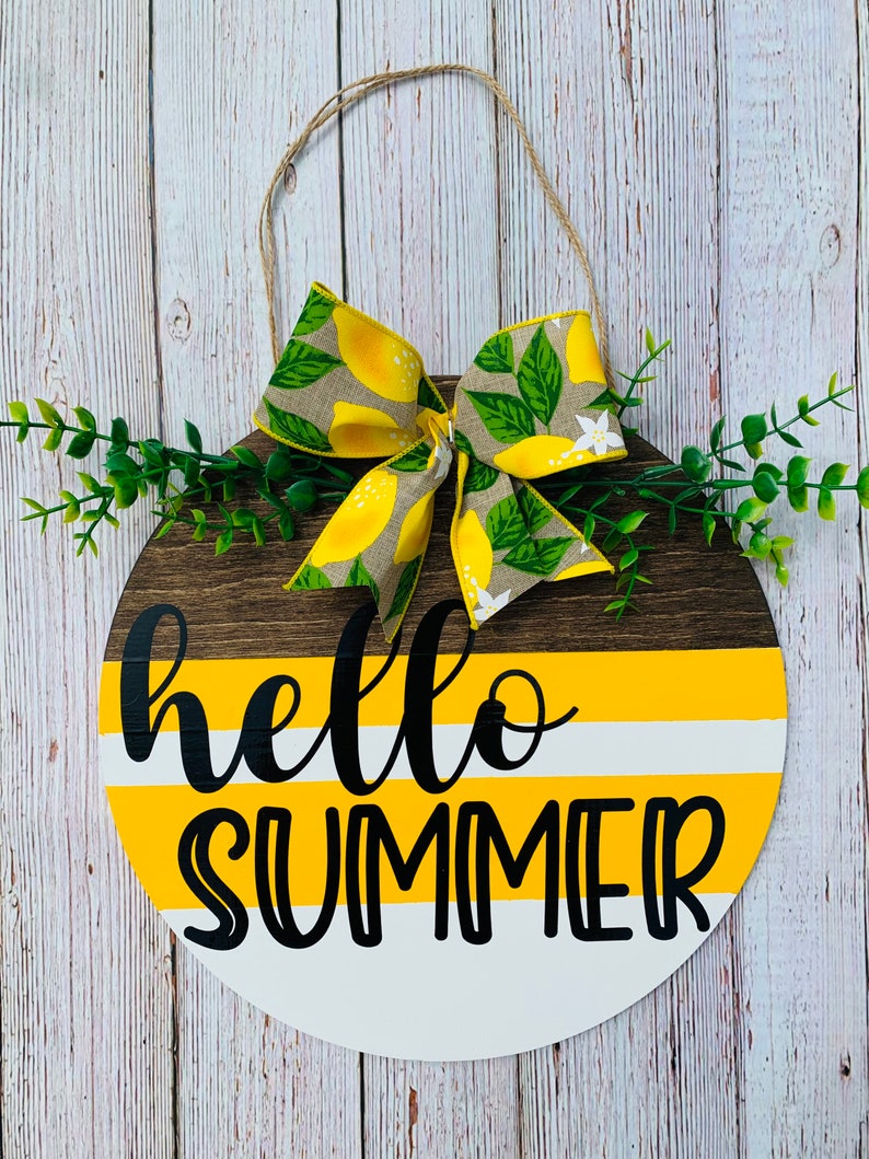 May include: A round wooden sign with a yellow and white striped background and the text "hello summer" in black. A yellow and green lemon print bow is attached to the top of the sign.