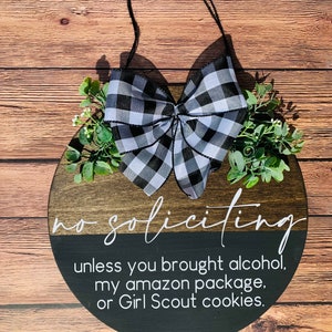Puede incluir: Un letrero redondo de madera con un lazo a cuadros blanco y negro y vegetaci&oacute;n. El letrero dice "no soliciting unless you brought alcohol, my amazon package, or Girl Scout cookies."