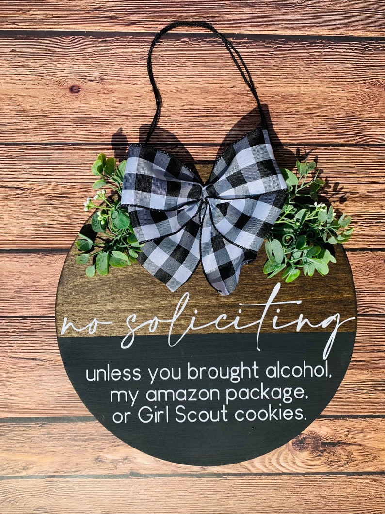Puede incluir: Un letrero redondo de madera con un lazo a cuadros blanco y negro y vegetaci&oacute;n. El letrero dice "no soliciting unless you brought alcohol, my amazon package, or Girl Scout cookies."