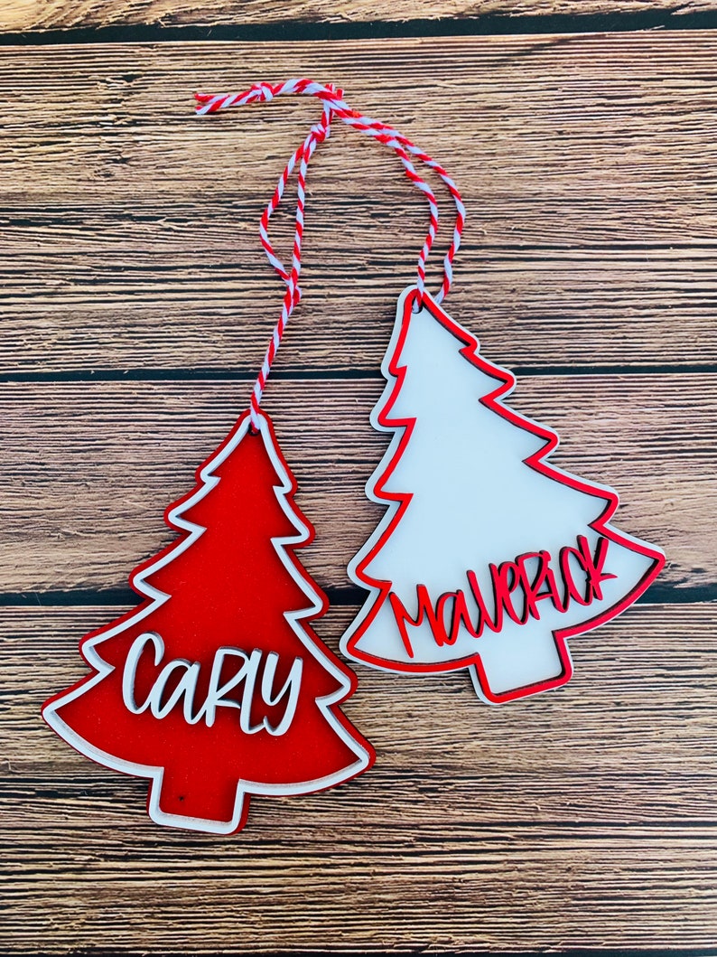 K&ouml;nnte beinhalten: Zwei rote und wei&szlig;e Weihnachtsbaumschmuckst&uuml;cke mit den Namen "Carly" und "Mallory" in wei&szlig; darauf.