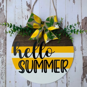 May include: A round wooden door hanger with a yellow and white striped background. The words "hello summer" are written in black cursive lettering. A lemon-themed bow is attached to the top of the hanger.