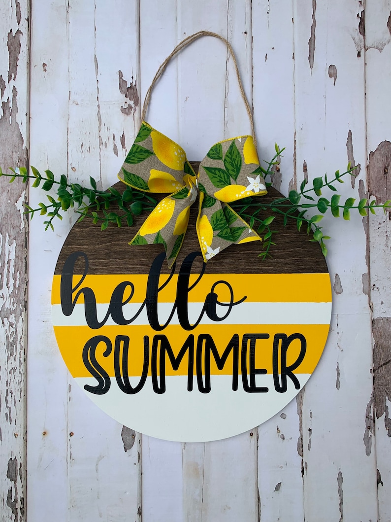 May include: A round wooden door hanger with a yellow and white striped background. The words "hello summer" are written in black cursive lettering. A lemon-themed bow is attached to the top of the hanger.