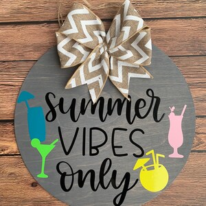 K&ouml;nnte beinhalten: Runder Holzschild mit dem Schriftzug "Summer Vibes Only" in schwarzer Schrift. Es zeigt farbenfrohe Cocktailgrafiken und eine dekorative Schleife mit Zickzackmuster. Das Schild ist grau mit einem Hintergrund aus nat&uuml;rlicher Holzmaserung.