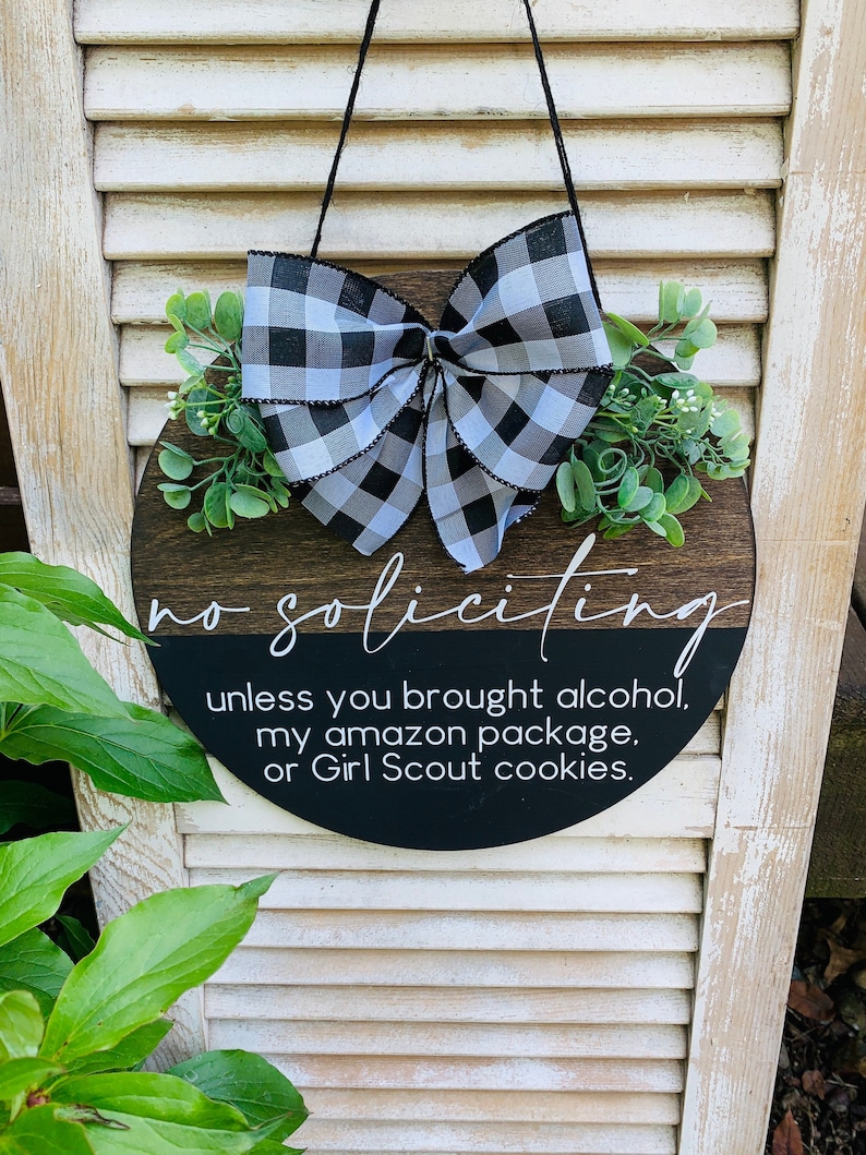 Puede incluir: Un letrero redondo de madera con un lazo a cuadros blanco y negro y vegetaci&oacute;n. El letrero dice "no soliciting unless you brought alcohol, my amazon package, or Girl Scout cookies."