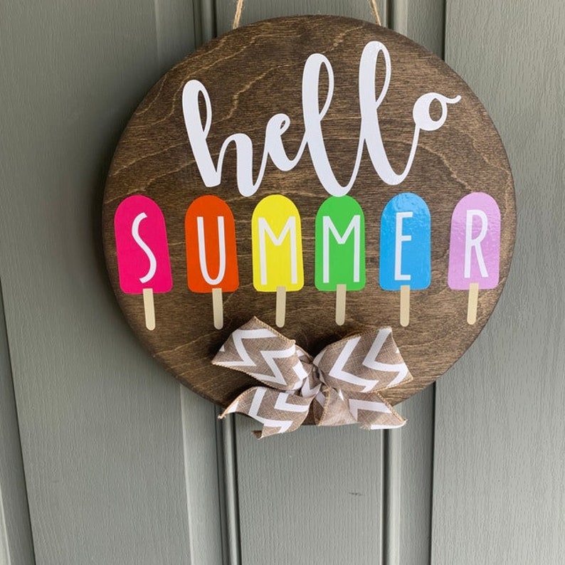 May include: A round wooden sign with the text "hello summer" in white lettering. The sign is decorated with colorful popsicle shapes with each letter of the word "summer" on a different popsicle.