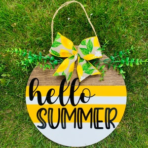 May include: A round wooden sign with a yellow and white striped background. The sign says "hello summer" in black lettering. A lemon-patterned bow is attached to the top of the sign.