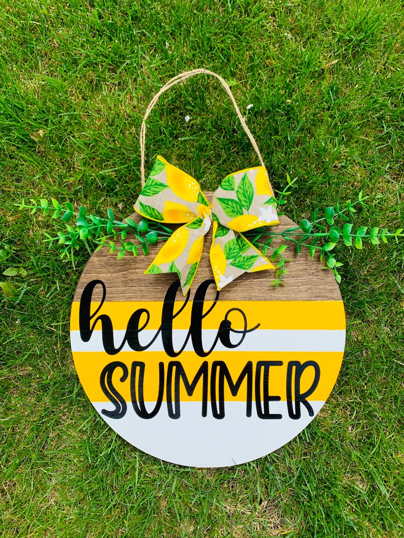 May include: A round wooden sign with a yellow and white striped background. The sign says "hello summer" in black lettering. A lemon-patterned bow is attached to the top of the sign.