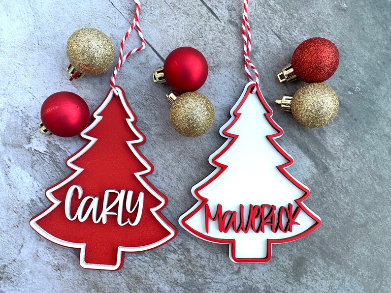 K&ouml;nnte beinhalten: Zwei rot-wei&szlig;e Weihnachtsbaumschmuckst&uuml;cke mit den Namen "Carly" und "Maverick" darauf. Die Schmuckst&uuml;cke h&auml;ngen an einer rot-wei&szlig;en Schnur.