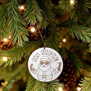 May include: A circular wooden Christmas tree decoration featuring the words "SIX SEVEN" and a Santa Claus illustration wearing sunglasses. The numbers "6" and "1" are on either side of Santa. The ornament hangs from a brown string against a Christmas tree.