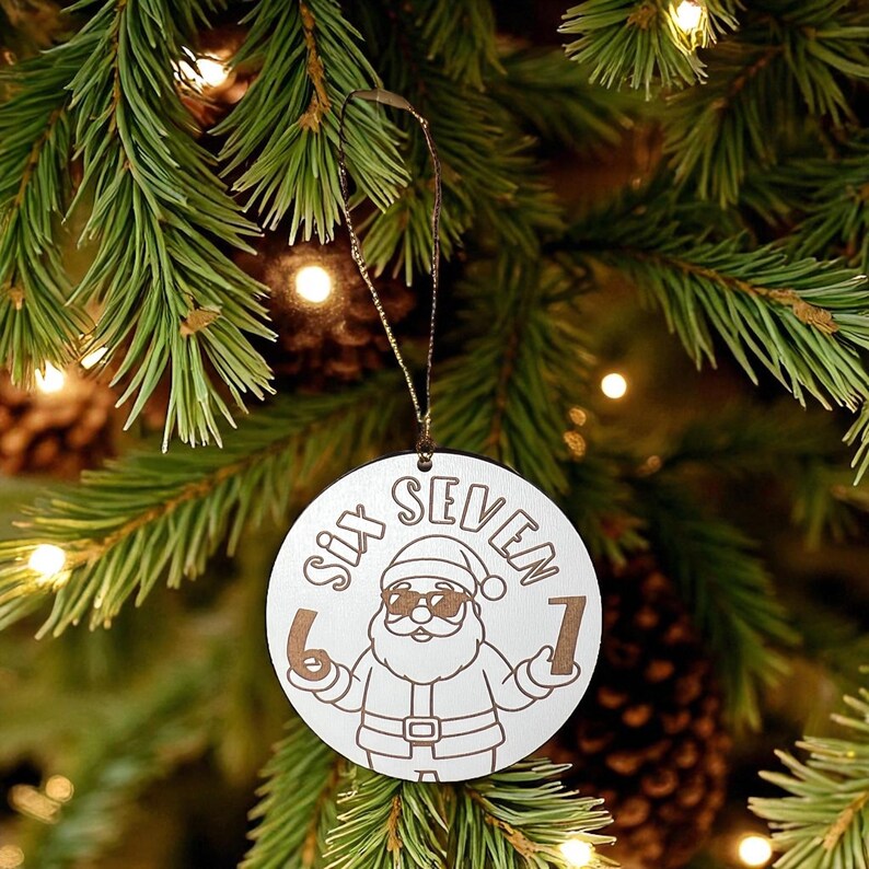 May include: A circular wooden Christmas tree decoration featuring the words "SIX SEVEN" and a Santa Claus illustration wearing sunglasses. The numbers "6" and "1" are on either side of Santa. The ornament hangs from a brown string against a Christmas tree.