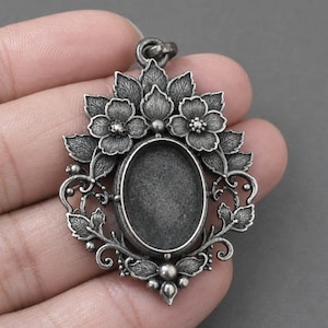 Puede incluir: Un colgante de metal en tono plateado envejecido con un engaste ovalado central rodeado de intrincados motivos florales y de hojas. El marco decorativo incluye pétalos detallados, follaje y pequeños acentos esféricos con una anilla superior.
