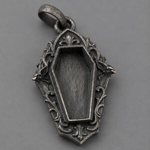 Puede incluir: Colgante de plata con dise&ntilde;o de ata&uacute;d. El colgante presenta una forma de ata&uacute;d abierto dentro de un marco ornamentado de estilo g&oacute;tico. El marco tiene adornos decorativos y un lazo para una cadena.