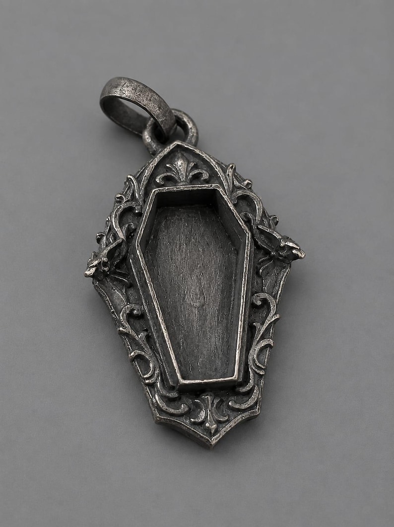 Puede incluir: Colgante de plata con dise&ntilde;o de ata&uacute;d. El colgante presenta una forma de ata&uacute;d abierto dentro de un marco ornamentado de estilo g&oacute;tico. El marco tiene adornos decorativos y un lazo para una cadena.