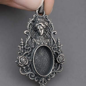 May include: An antique silver-toned metal pendant featuring an oval central bezel setting. The frame is decorated with intricate scrollwork, two sculpted roses on the sides, and a relief portrait of a person at the top with a lattice background.