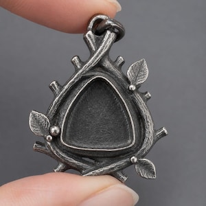 Puede incluir: Un colgante de metal en tono plateado oscuro con un engaste central triangular hundido. El marco está diseñado como ramas entrelazadas con pequeñas hojas texturizadas y diminutas esferas, rematado con una anilla circular.