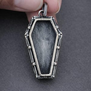 May include: A silver coffin pendant with a textured interior. The pendant has a rectangular shape with rounded edges, framed by a silver border and bamboo-like detailing. A bail is at the top for a chain.