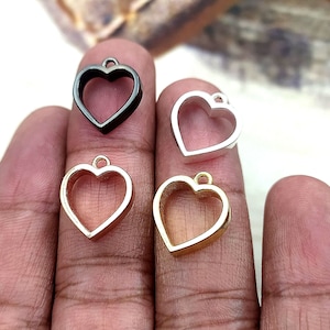 Könnte beinhalten: Vier herzförmige Metall-Anhänger mit einer Öse oben. Die Anhänger sind in Schwarz, Silber, Gold und Klar.