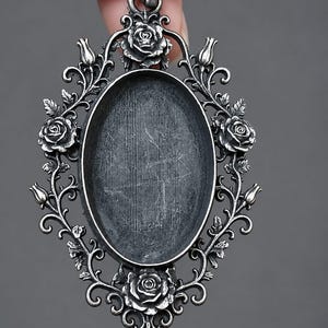 May include: An oval-shaped metal pendant tray with a dark, oxidised silver finish. The frame features intricate, raised floral detailing with sculpted roses and leaves surrounding a central, recessed oval area for a custom insert.