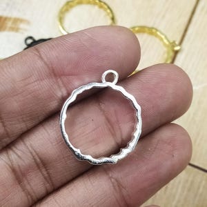 Pode incluir: Um pingente prateado em forma de círculo aberto com uma borda texturizada e ondulada. O pingente tem um pequeno laço na parte superior para prender a uma corrente ou outro componente de joia. Outros anéis dourados são visíveis no fundo.