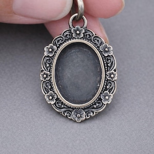 May include: Silver-toned oval pendant with a floral border. The pendant has an empty center, and a loop at the top for a chain. The floral design includes small flowers and swirling patterns. The pendant is held by a person's fingers.