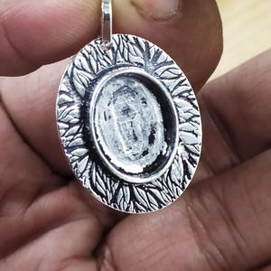 May include: Oval silver pendant with a leaf design around the bezel. The pendant has a loop at the top for a chain. The centre is a recessed area for a stone or other embellishment.