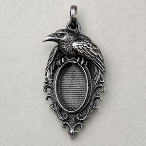 May include: A silver-toned metal pendant featuring a detailed raven perched atop an oval frame. The frame has a horizontal ribbed texture inside and is surrounded by ornate, scrolling metalwork. A circular bail is attached at the top.