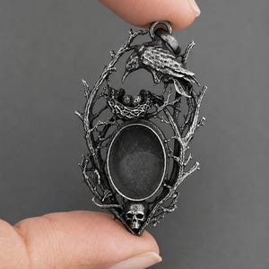 May include: An oval silver-toned metal pendant featuring a detailed bird perched above a nest containing two small eggs. The frame is composed of textured branches and twigs, with a small human skull positioned at the base of the central oval setting.