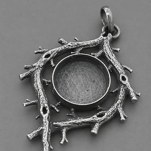Puede incluir: Un colgante de metal plateado con un engaste circular en el centro, rodeado por un marco texturizado que imita la apariencia de ramas de árbol entrelazadas. La pieza incluye una pequeña anilla en la parte superior para su sujeción.