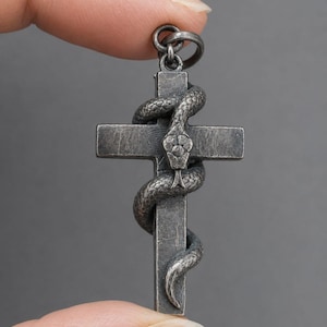 May include: A dark, oxidised silver-toned metal cross pendant featuring a textured snake coiled around the vertical and horizontal bars. The snake's head is positioned at the centre of the cross, and a small circular jump ring is attached at the top.