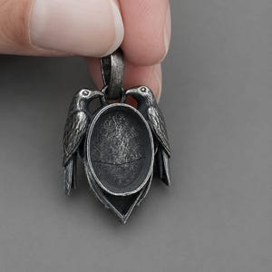 Op de afbeelding: Zilverkleurige hanger met twee gedetailleerde ravenfiguren die een ovale zetting flankeren. De hanger heeft een getextureerde, antieke afwerking en is opgehangen aan een kleine lus. Het ontwerp is symmetrisch, op een grijze achtergrond.