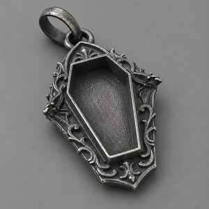 Puede incluir: Colgante plateado con un dise&ntilde;o de ata&uacute;d dentro de un marco ornamentado de estilo g&oacute;tico. El colgante tiene una anilla para una cadena y est&aacute; sobre un fondo gris. El ata&uacute;d est&aacute; vac&iacute;o.