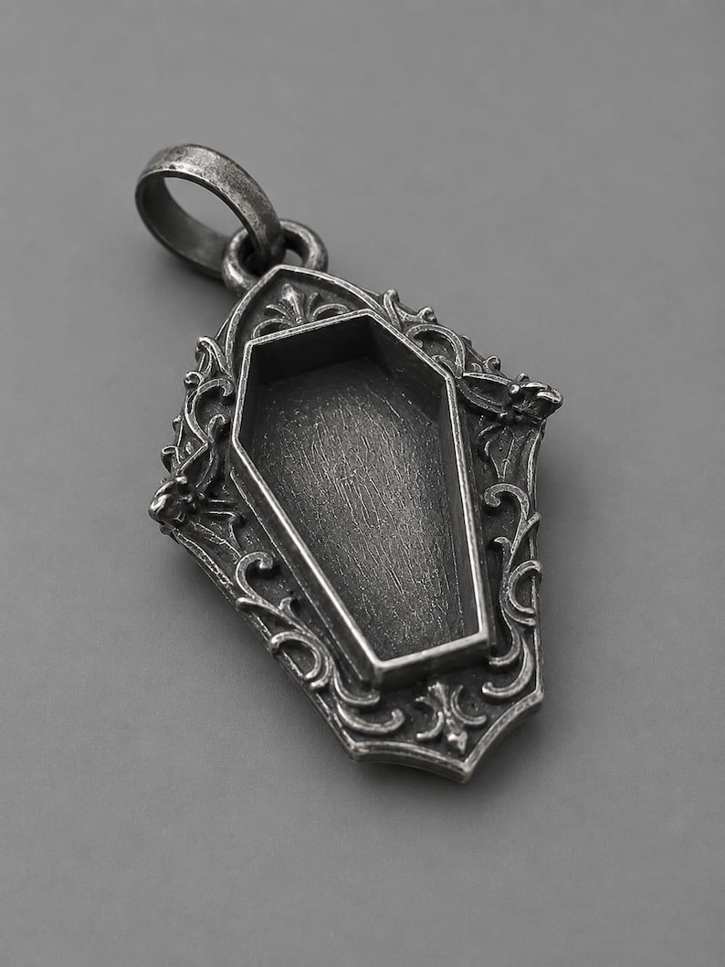 Puede incluir: Colgante plateado con un dise&ntilde;o de ata&uacute;d dentro de un marco ornamentado de estilo g&oacute;tico. El colgante tiene una anilla para una cadena y est&aacute; sobre un fondo gris. El ata&uacute;d est&aacute; vac&iacute;o.