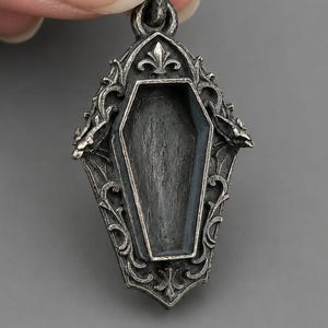 Puede incluir: Colgante plateado con dise&ntilde;o de ata&uacute;d. El colgante tiene un marco ornamentado de estilo g&oacute;tico con volutas y una flor de lis en la parte superior. El ata&uacute;d est&aacute; vac&iacute;o y tiene una superficie texturizada.