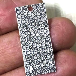 Può includere: Un piccolo ciondolo rettangolare argentato con un motivo organico strutturato. Il ciondolo ha un piccolo foro nella parte superiore per l'infilatura. La superficie è dettagliata con un design irregolare e ripetitivo.