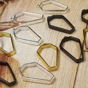 May include: A collection of coffin-shaped metal charms in silver, gold, and black. These small, open-frame pendants are designed for jewelry making or crafting. The charms are scattered on a light brown wooden surface.