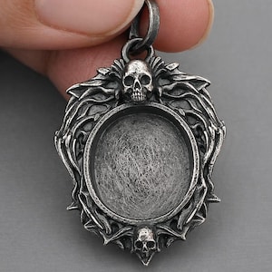 May include: A silver-toned pendant with a skull and ornate, gothic-style detailing. The pendant has a circular recessed area, possibly for a stone or other insert. The design includes two additional skulls and a decorative border.