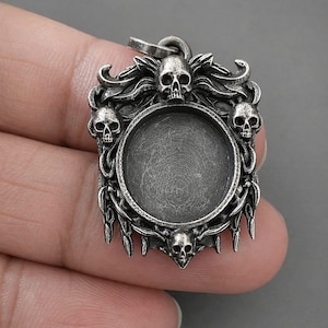 Op de afbeelding: Zilverkleurige hanger met een centrale ronde setting, omringd door sierlijke details en schedelmotieven. De hanger heeft een oogje om aan een ketting te bevestigen. Het ontwerp is gotisch en macaber.