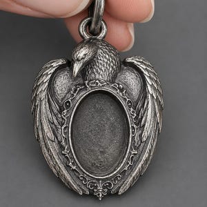 May include: A silver-toned metal pendant featuring a detailed raven with spread wings. The center of the pendant contains an oval-shaped recessed frame with an ornate, decorative border. The piece includes a circular bail at the top for hanging.