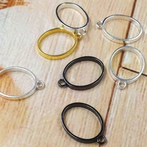 以下が含まれることがあります： 銀、金、黒の楕円形メタルリングブランクの詰め合わせ。各リングには、チャームなどを取り付けるための小さなループが付いています。リングは、明るい茶色の木製の表面に表示されています。