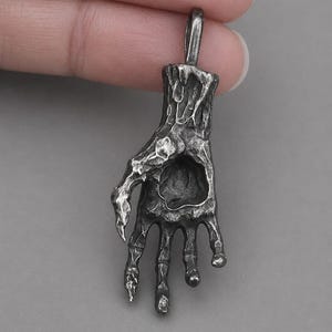 May include: A silver-toned pendant in the shape of a skeletal hand. The pendant has a textured, weathered appearance with intricate details. The hand is depicted with elongated fingers and a loop for a chain.
