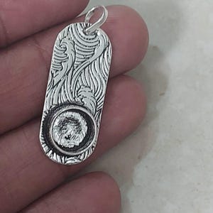 以下が含まれることがあります： 彫刻デザインのシルバーペンダント。ペンダントは、丸みを帯びたエッジと小さなループを備えた長方形の形状をしています。デザインには、渦巻き模様と詳細な画像を含む円形エンブレムが含まれています。