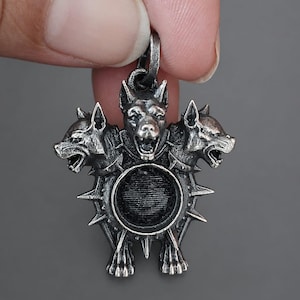 May include: A silver pendant depicting a three-headed dog. The pendant has a circular central feature and is encircled by spikes. The dog heads are detailed with open mouths and pointed ears. The pendant is held by a hand.