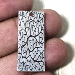 Può includere: Ciondolo rettangolare in argento con un motivo a foglie dettagliato. Il ciondolo ha un piccolo foro nella parte superiore per una catena. Il design presenta una serie di forme di foglie interconnesse, creando una superficie strutturata.